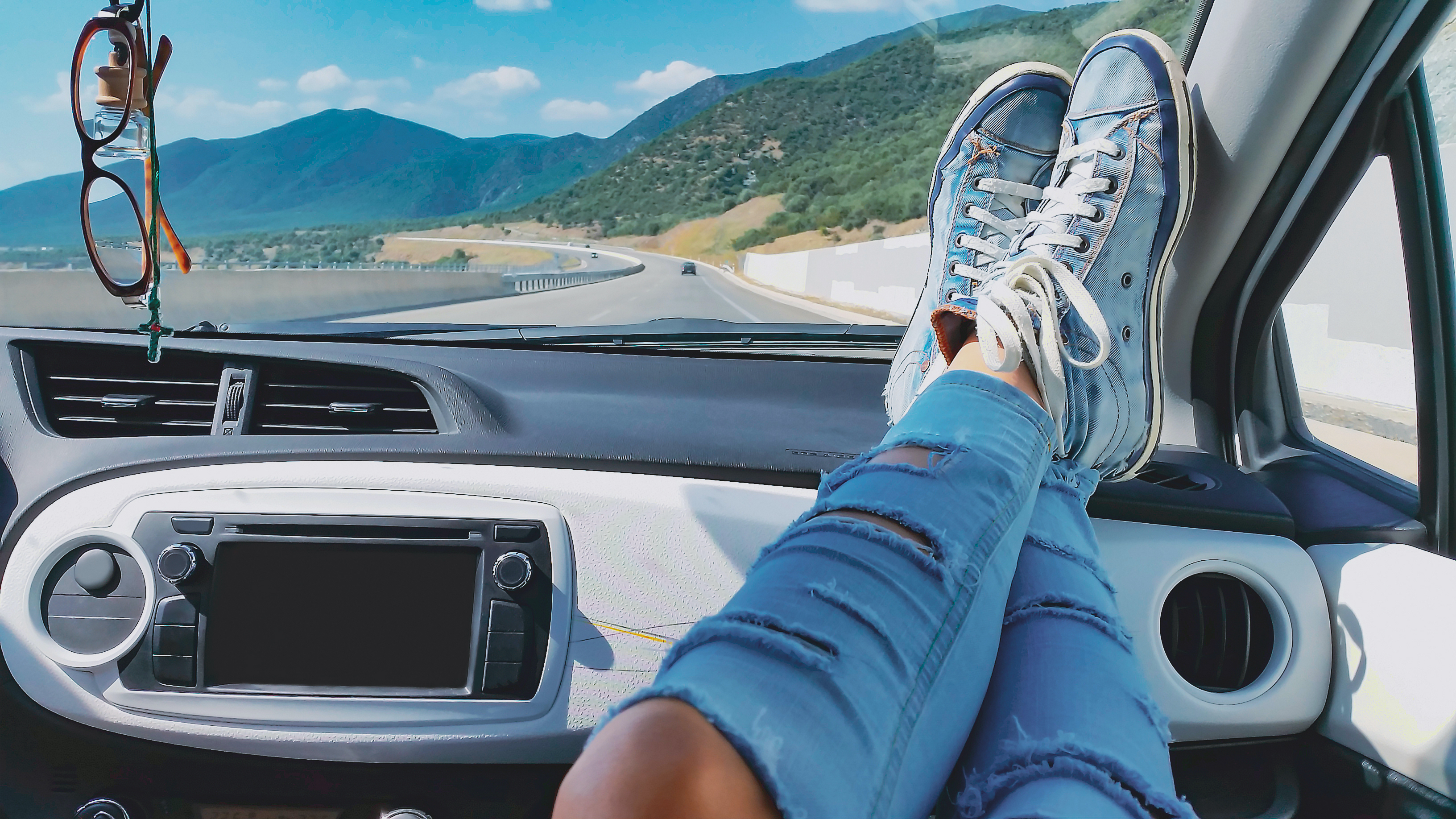 Female legs in ripped jeans and blue sneakers inside car with mountains landscape at background. Woman relaxing during road trip