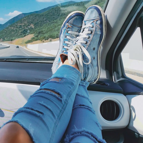 Female legs in ripped jeans and blue sneakers inside car with mountains landscape at background. Woman relaxing during road trip